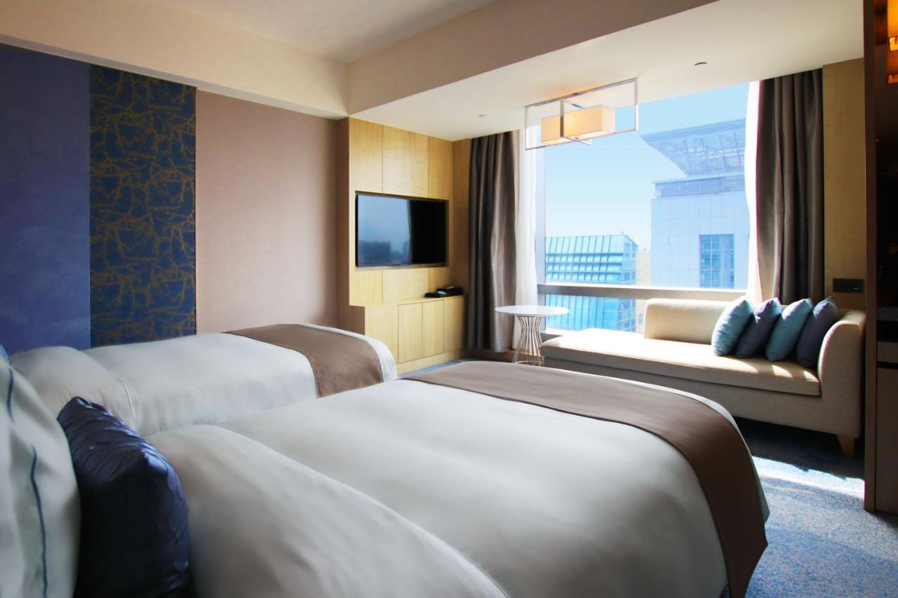 Caesar Park Hotel Banqiao Taipei Exterior photo The photo shows a modern hotel room featuring two beds with white linens and a brown accent. There's a decorative wall with textured elements, and a flat-screen TV is mounted on one side. A large window allows natural light to flood the space, offeri