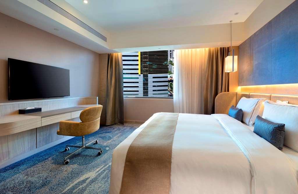 Caesar Park Hotel Banqiao Taipei Room photo The photo shows a modern hotel room featuring a large bed with white and beige bedding. There is a comfortable chair by a desk that includes a flat-screen TV mounted on the wall. The room has large windows allowing natural light to enter, with sheer 
