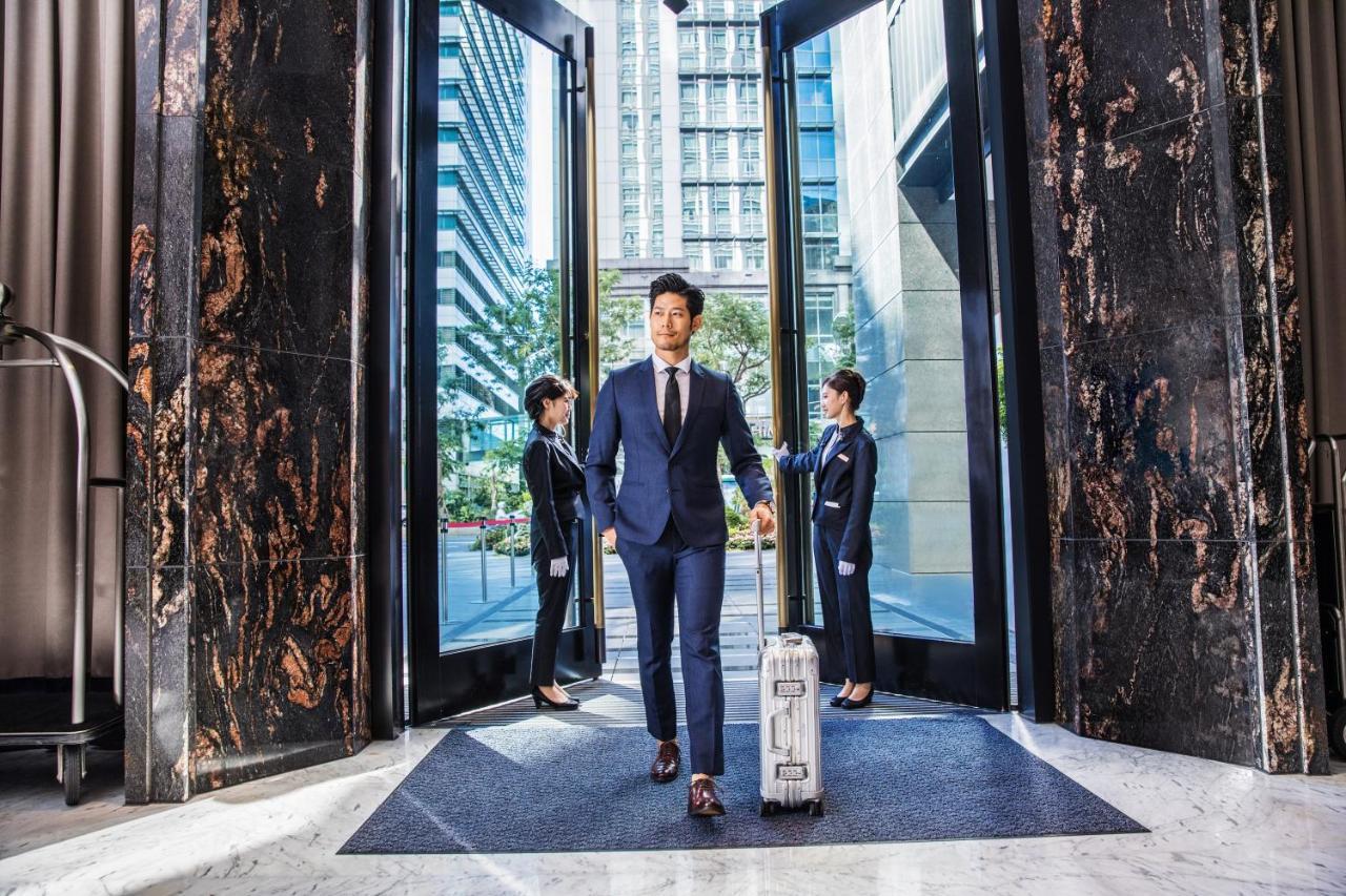 Caesar Park Hotel Banqiao Taipei Exterior photo The photo shows a professional setting, likely in a hotel or high-rise building. In the foreground, a man is walking through large glass doors, dressed in a tailored suit and carrying a suitcase. He appears confident and is the focal point of the sce