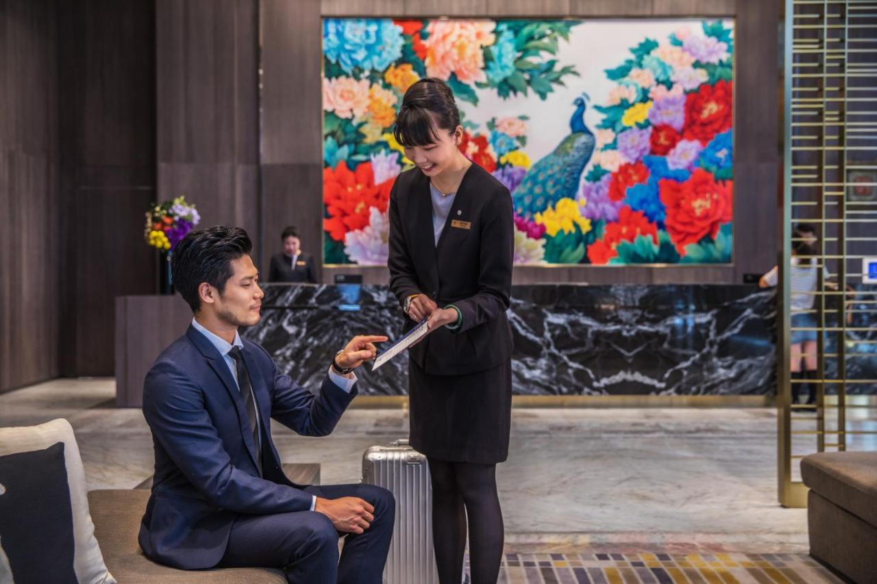 Caesar Park Hotel Banqiao Taipei Exterior photo The photo depicts a hotel lobby scene. In the foreground, a man in a tailored suit sits on a modern sofa, while a female staff member in a professional outfit is standing next to him, pointing at a tablet or document. The background features a vibran