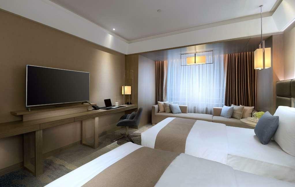 Caesar Park Hotel Banqiao Taipei Exterior photo The photo shows a modern hotel room featuring two beds with brown and white bedding. Against one wall, there's a flat-screen TV mounted on a wooden console. To the side, there's a desk with a chair, likely for working. Large windows are covered with 