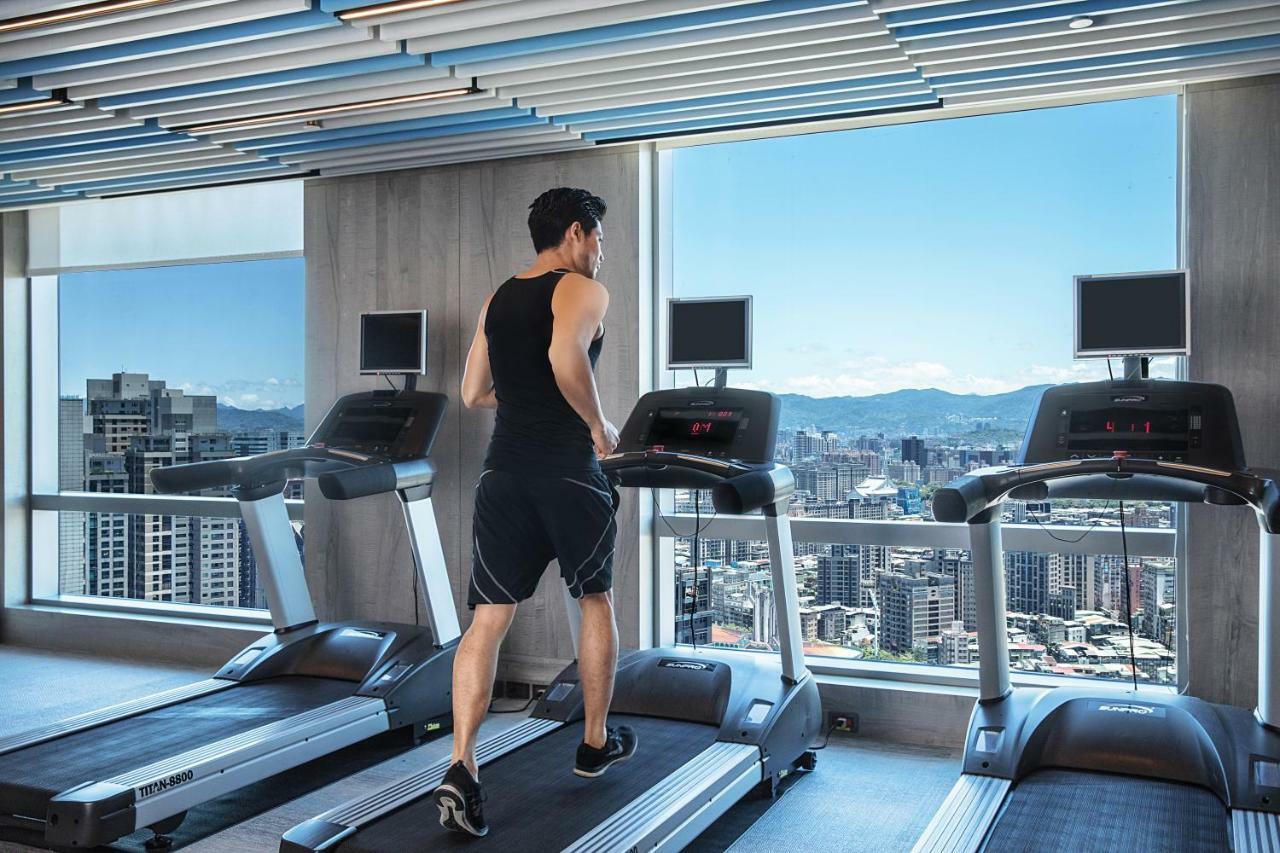 Caesar Park Hotel Banqiao Taipei Exterior photo The photo depicts a man running on a treadmill in a modern gym. He is wearing a sleeveless shirt and shorts, and appears to be focused on his workout. The gym has large windows that offer a panoramic view of a city skyline with distant mountains. The